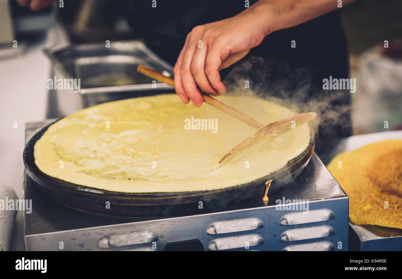 Realizzazione di Crepes Pancake nel mercato aperto festival fiera. una mano è rendere crepes all'aperto su una griglia metallica con bastone di legno su un outdoor summer fest Foto Stock