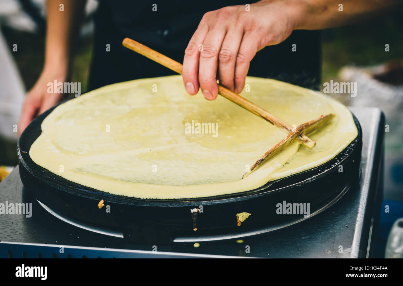 Realizzazione di Crepes Pancake nel mercato aperto festival fiera. una mano è rendere crepes all'aperto su una griglia metallica con bastone di legno su un outdoor summer fest Foto Stock