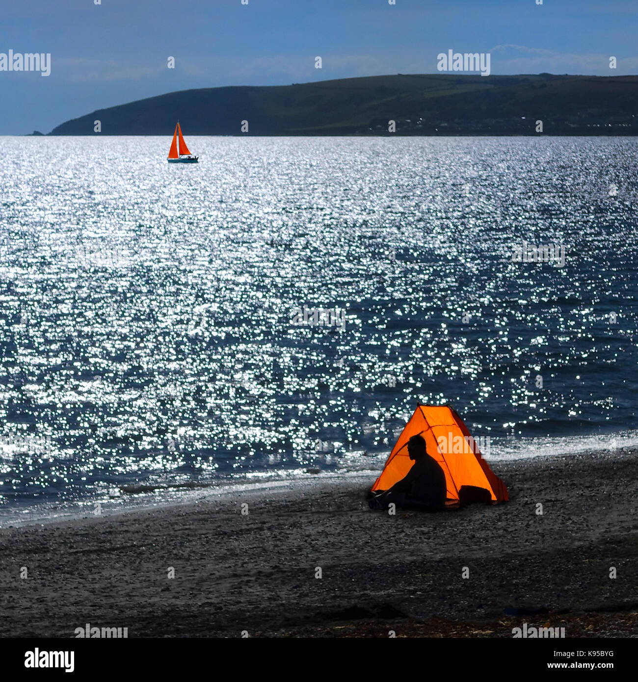 Stagliano uomo in arancione tenda sulla spiaggia della Cornovaglia con vele arancione in background Foto Stock