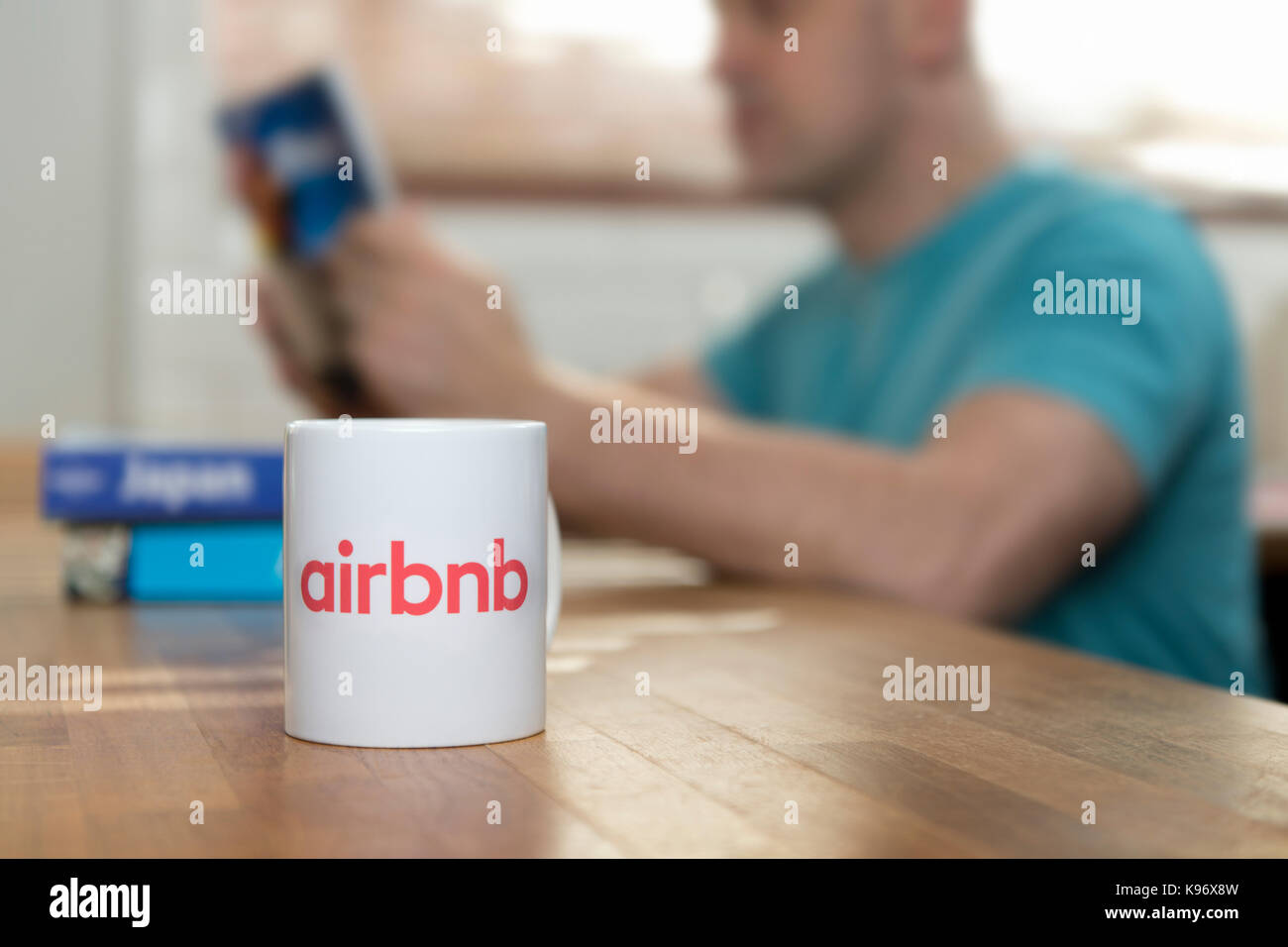 Un uomo legge guida viaggi libri in un appartamento che ha affittato usando le app Airbnb, e per i prodotti di marca Airbnb mug siede in primo piano (solo uso editoriale). Foto Stock