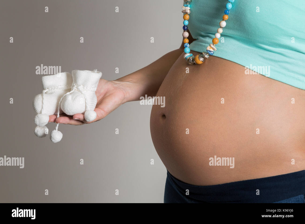 Donna incinta che mostra la sua pancia con una coppia di bianco piccolo bambino babbucce nella sua mano. Foto Stock