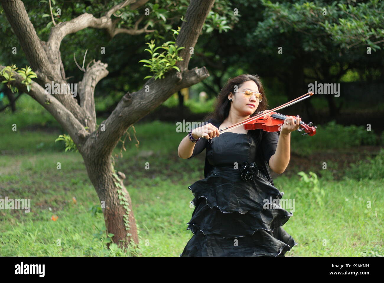 Una bella ragazza indiana musicista in abito nero riproduzione di un violino rosso nella parte anteriore di un albero e la natura Foto Stock