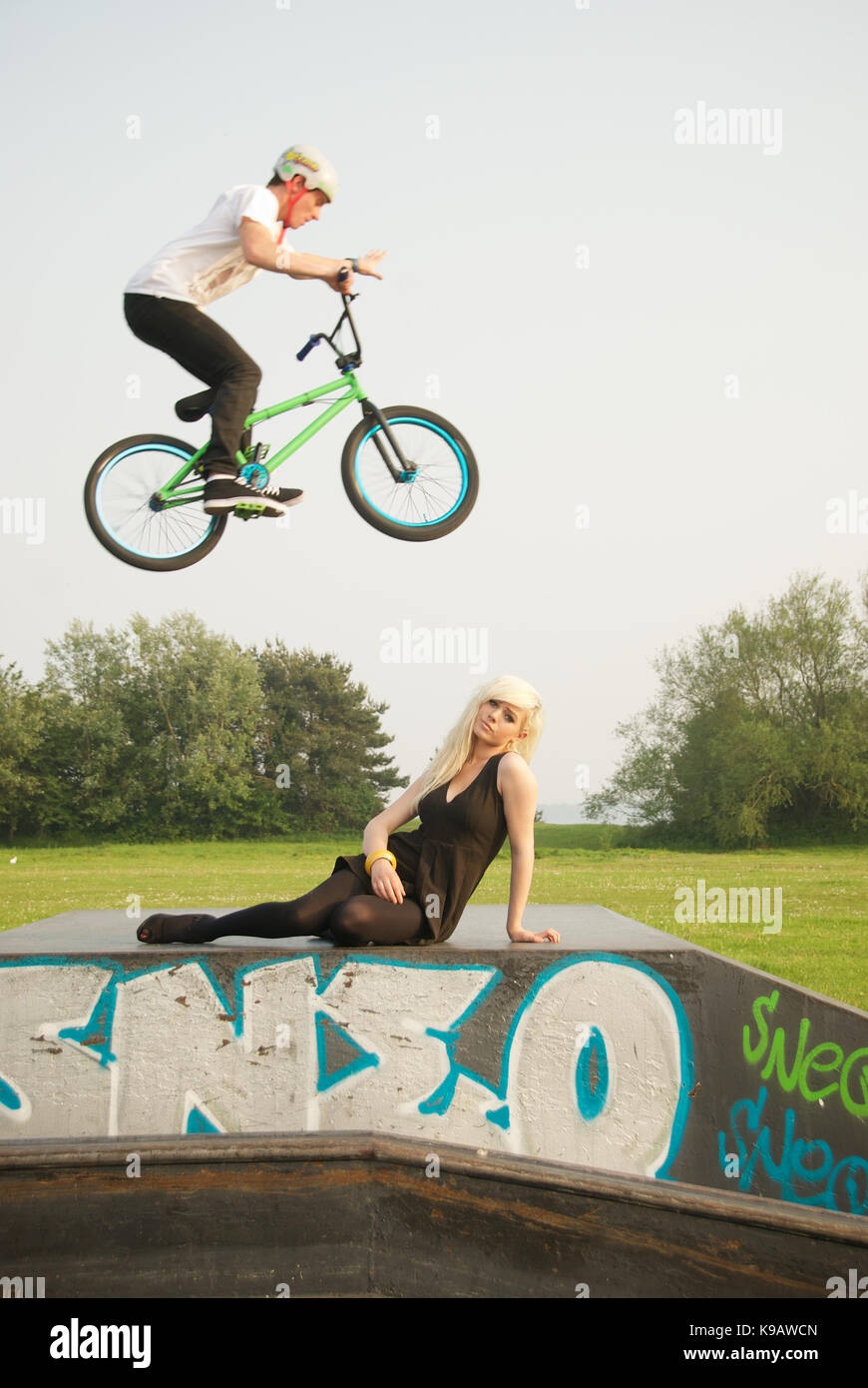 Bella ragazza bionda con stunt ciclista airborne al di sopra di lei in un parco Foto Stock