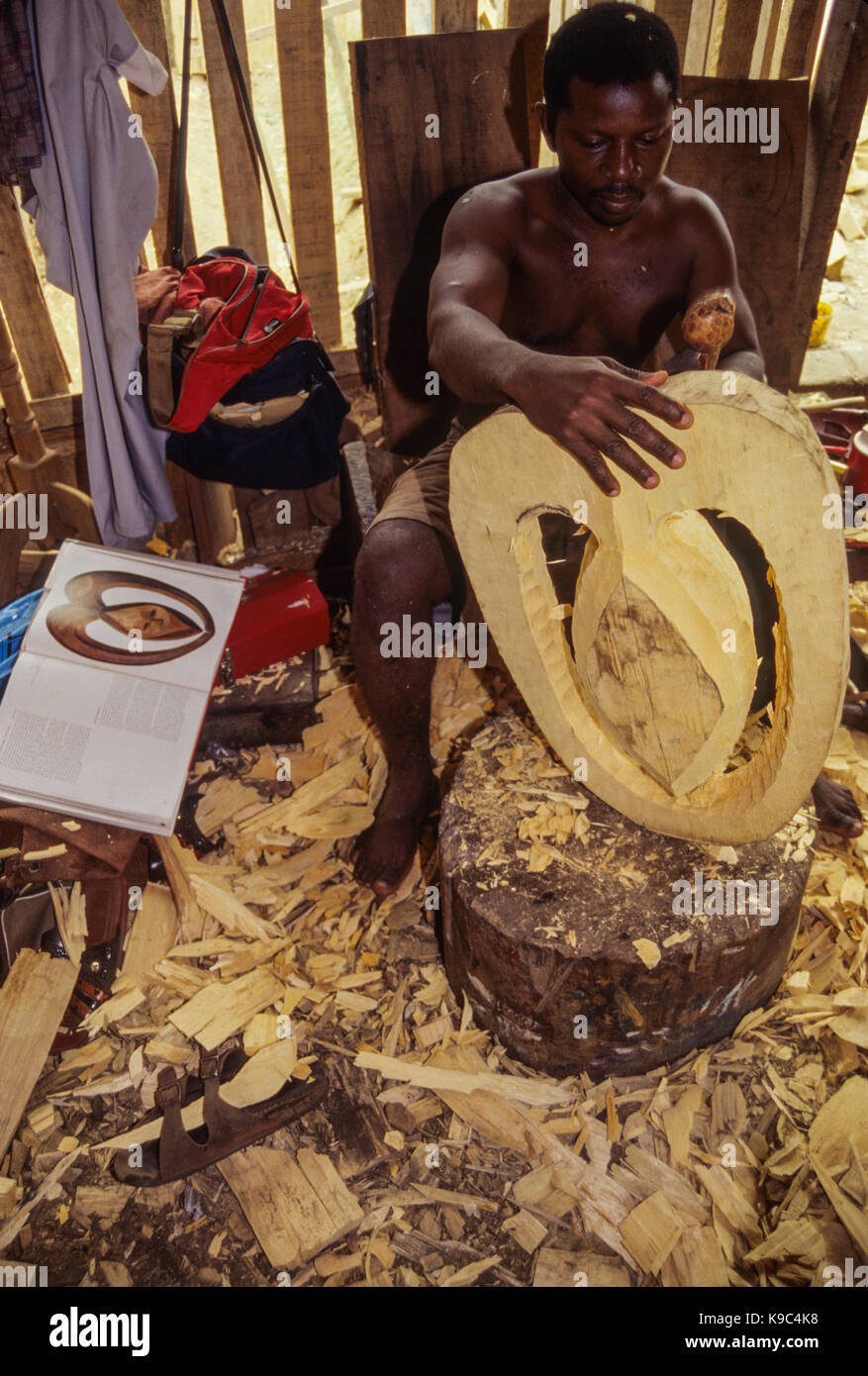 Abidjan, Costa d'Avorio, Costa d'Avorio. Utilizzando un'arte africana libro per fare riferimento a questo ghanesi Carver rende una replica di una maschera Kwele, a persone che vivono alle frontiere del Camerun, il Gabon e il Congo. Foto Stock