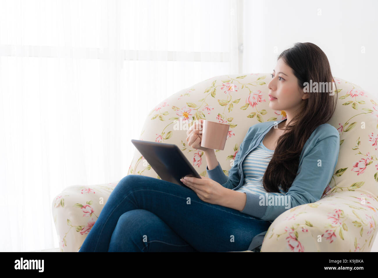 Piacevole donna attraente coppa di ritegno mug guardando fuori della finestra visualizza e seduto sul divano godendo di tempo di vacanza con la pastiglia mobile computer. Foto Stock
