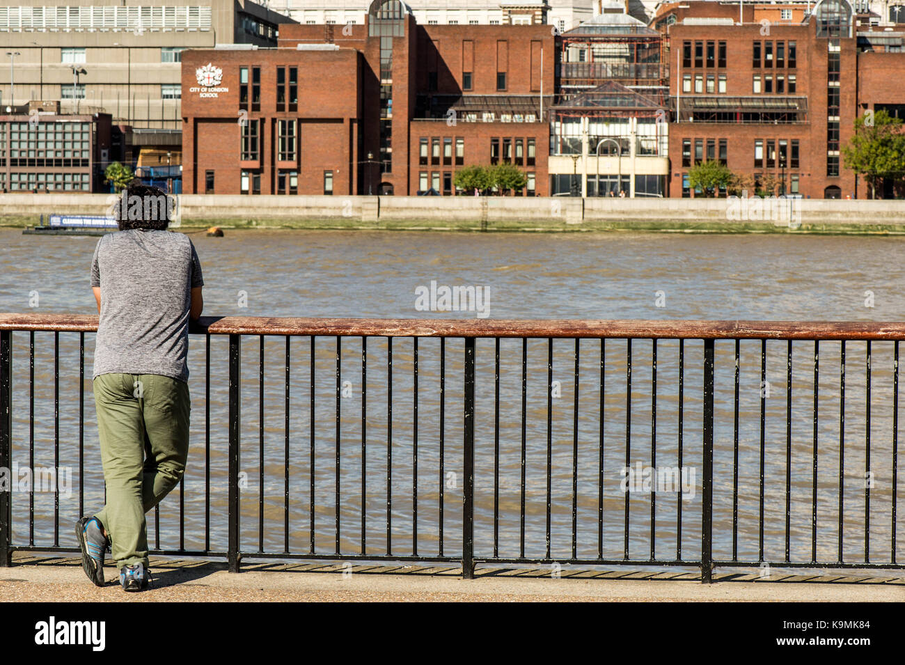 L'uomo con il suo ritorno alla telecamera appoggiato su di una recinzione di sicurezza guardando attraverso il fiume Tamigi a Londra Regno unito in un ragionato releaxd pongono, usura Foto Stock