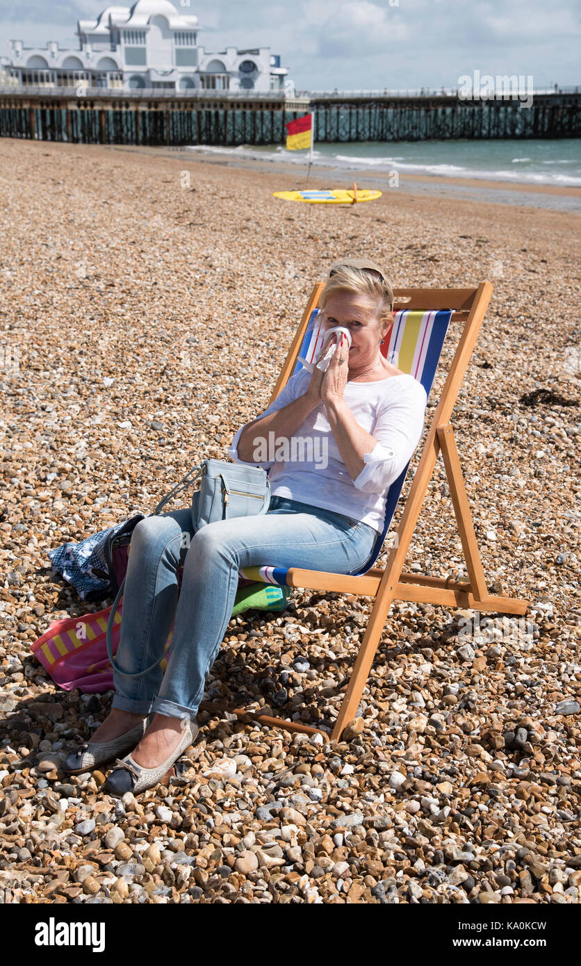 Donna seduta in una sedia a sdraio in riva al mare che soffre di un naso che cola. soffia il naso Foto Stock