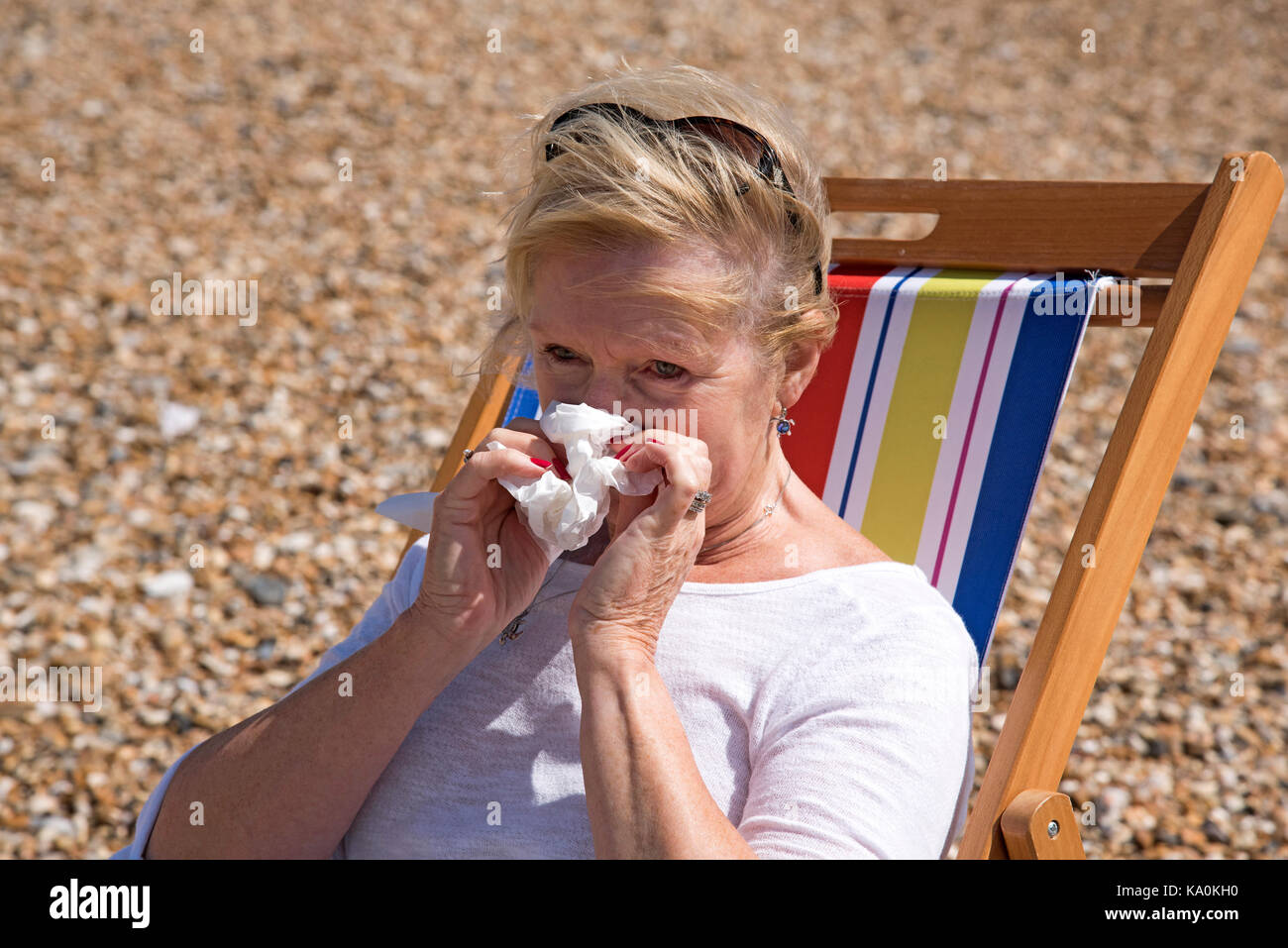 Donna seduta in una sedia a sdraio in riva al mare che soffre di un naso che cola. soffia il naso Foto Stock