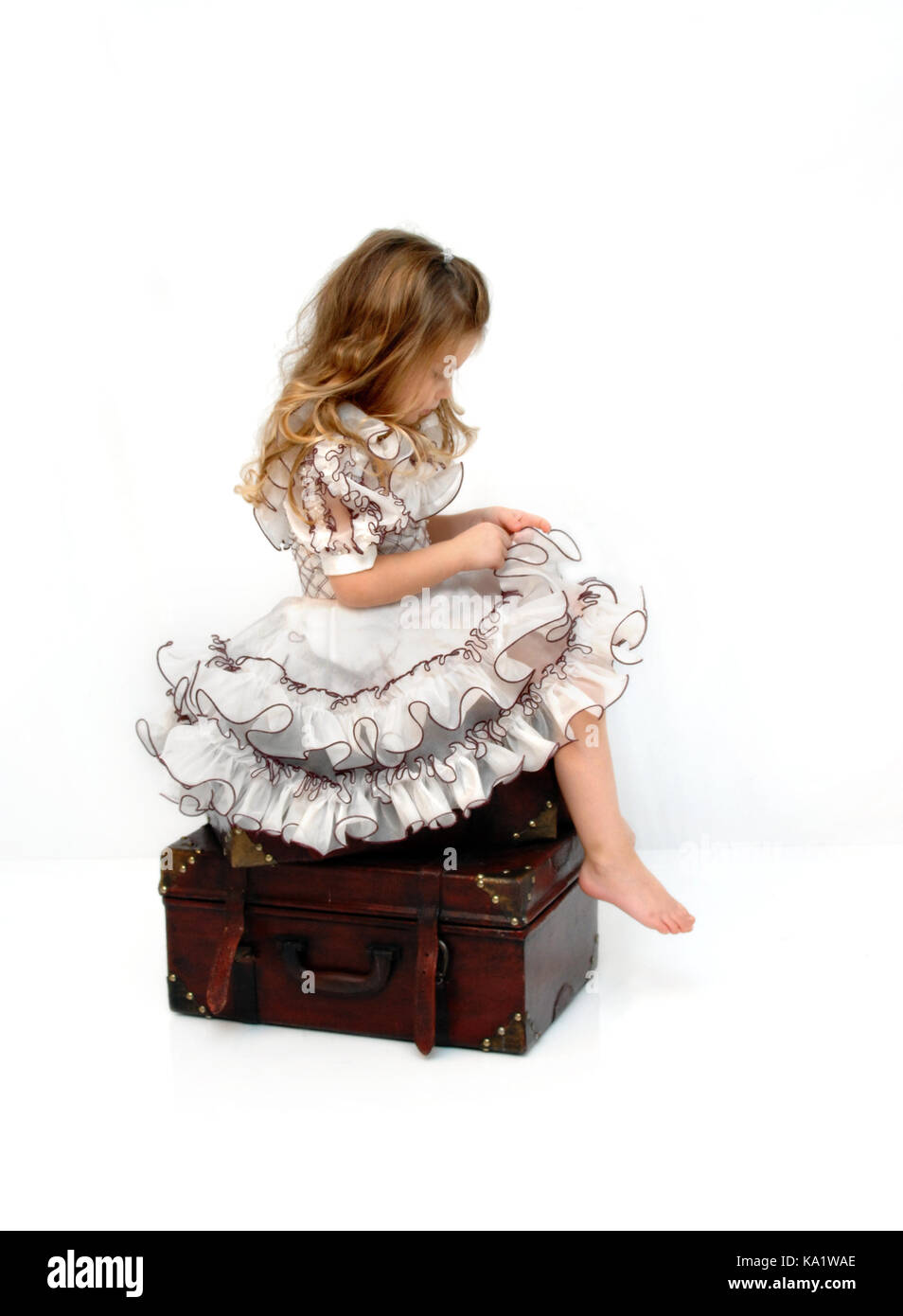 Splendida bambina si siede su una pila di valigie in legno. indossa un abito pagent e guardando verso il basso perso nel pensiero. Foto Stock