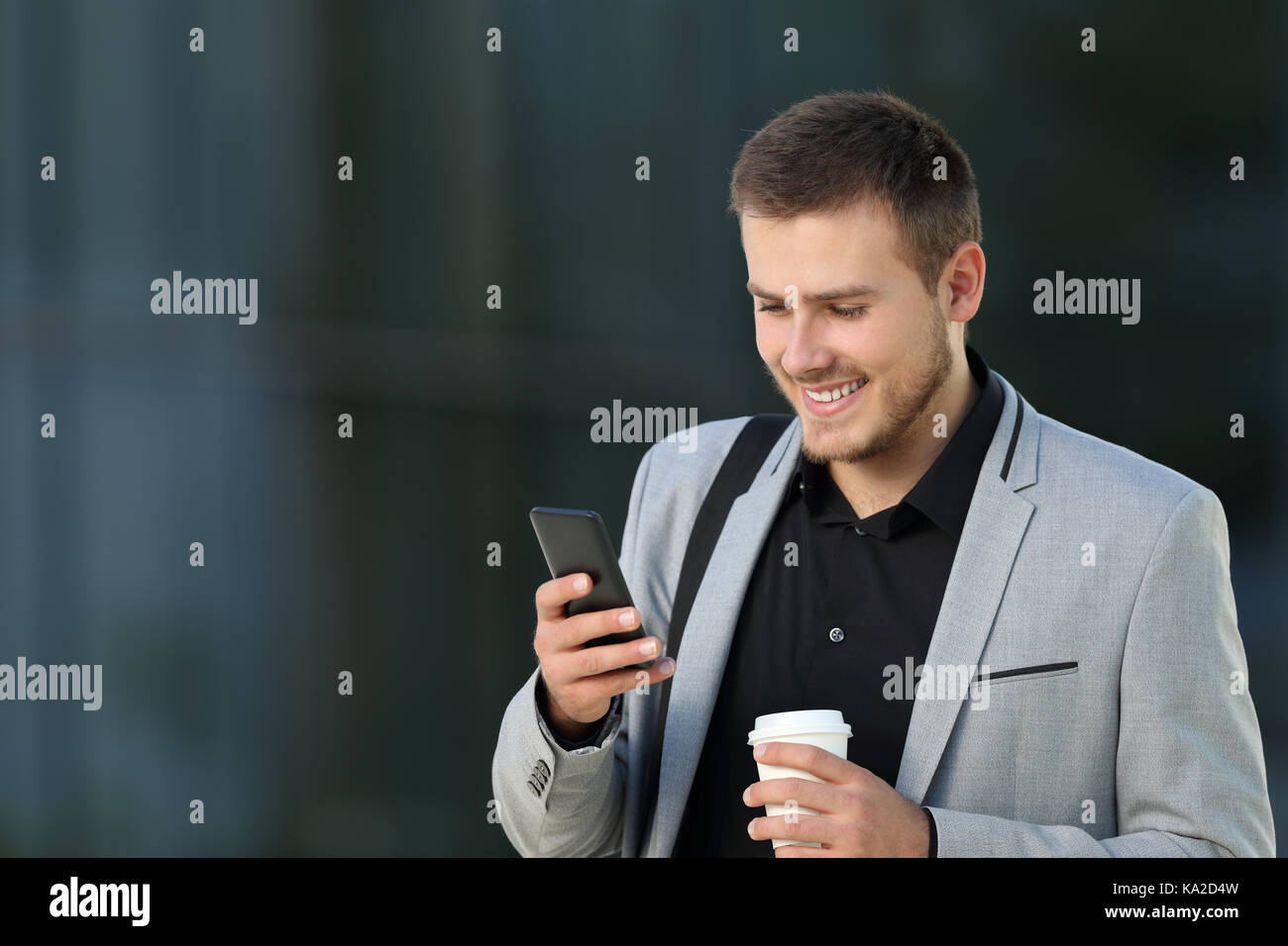 Una felice lettura esecutivo un telefono cellulare messaggio e tenendo un take away drink camminando sulla strada Foto Stock