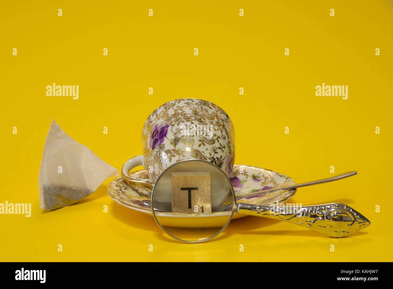 Bustine di the con tazza e piattino e cucchiaio d'argento con la lettera t piastrella ingrandito con lente di ingrandimento lente su sfondo giallo. amore tea ancora il concetto di vita Foto Stock