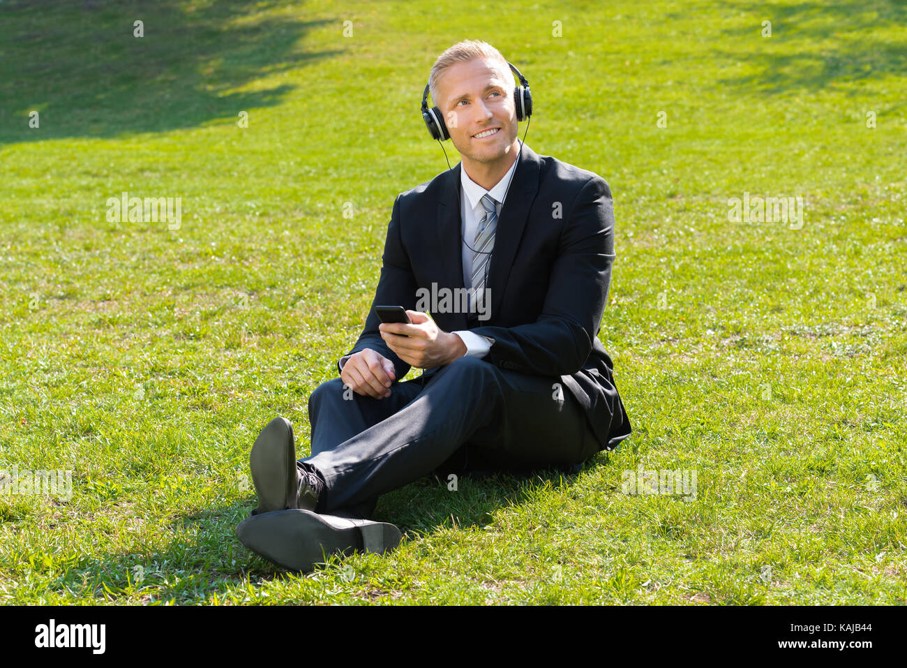 Giovane Imprenditore sorridente seduto sull'erba ascoltando musica in posizione di parcheggio Foto Stock