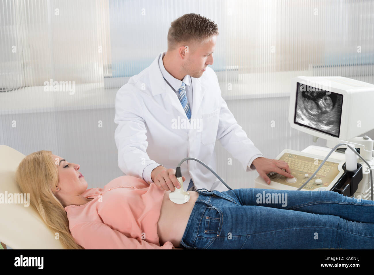 Medico maschio muovendo il trasduttore di ultrasuoni sulla donna incinta la pancia mentre guardando lo schermo in ospedale Foto Stock
