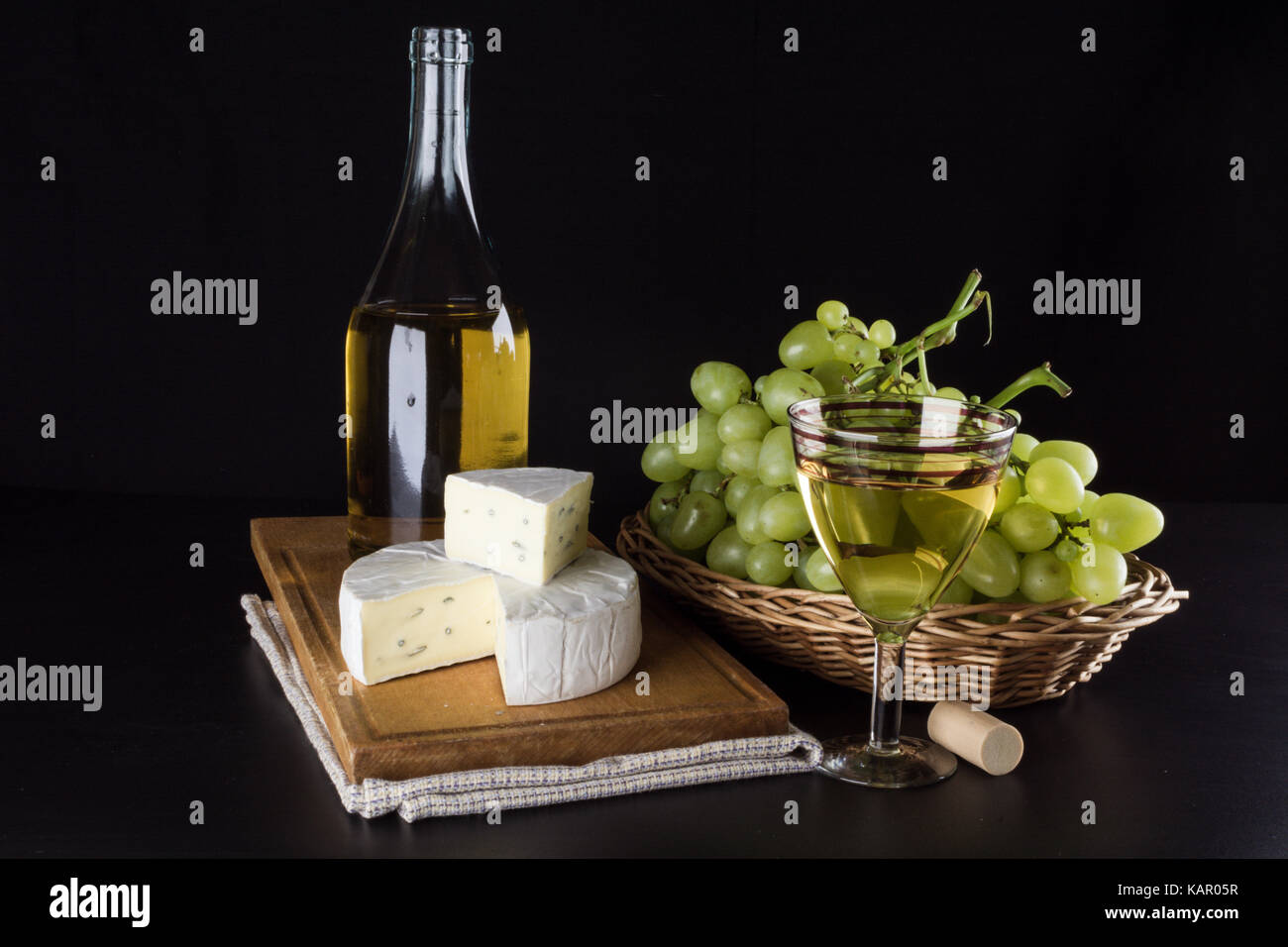 Una bottiglia di vino bianco in background, uva in un canestro e il formaggio dor blu e un bicchiere di vino bianco su sfondo nero Foto Stock