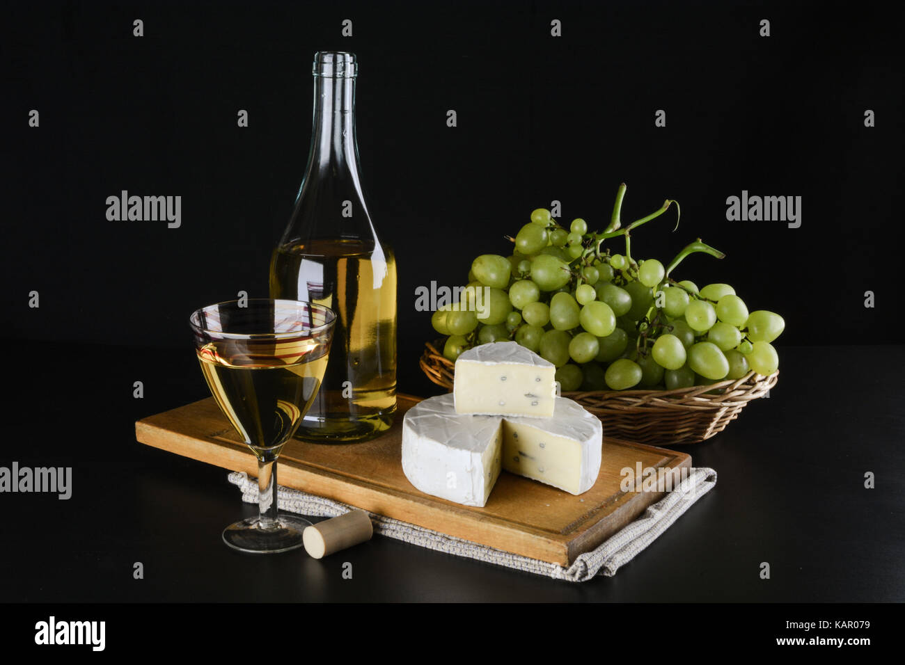 Una bottiglia di vino bianco in background, uva in un canestro e il formaggio dor blu e un bicchiere di vino bianco su sfondo nero Foto Stock
