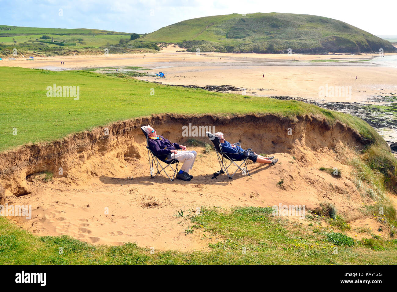 Uomo maturo e la donna riparo dal vento nelle dune di sabbia mentre crogiolandovi sotto i raggi del sole all'Daymer Bay, Cornwall, England, Regno Unito Foto Stock