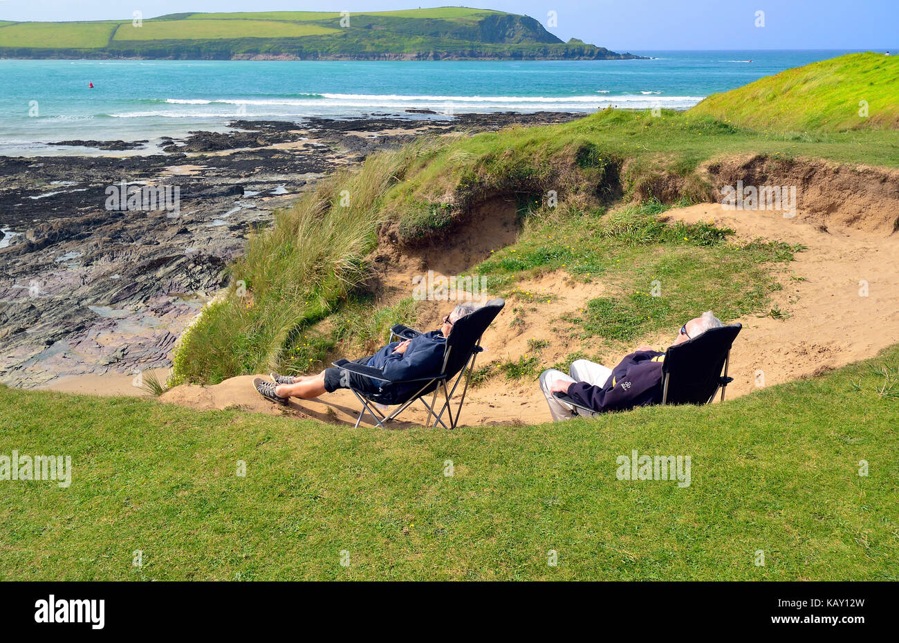 Uomo maturo e la donna riparo dal vento nelle dune di sabbia mentre crogiolandovi sotto i raggi del sole all'Daymer Bay, Cornwall, England, Regno Unito Foto Stock