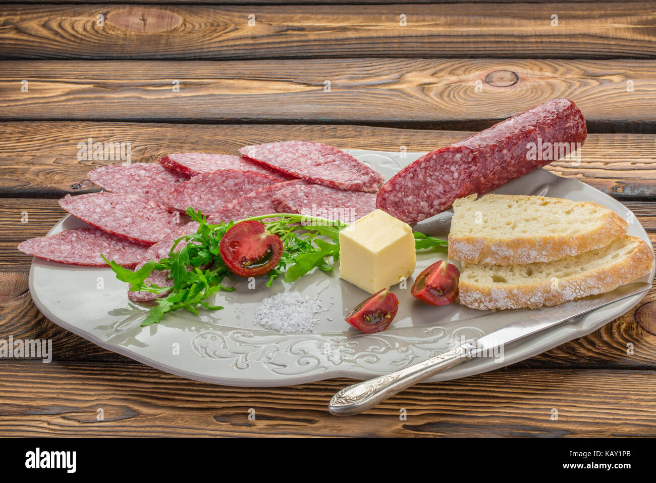 Materie salsiccia affumicata salame sulla piastra bianca, coltello. Ingredienti per i panini con salsiccia timo, erbe, pomodori, pane bianco, il burro sul backg in legno Foto Stock