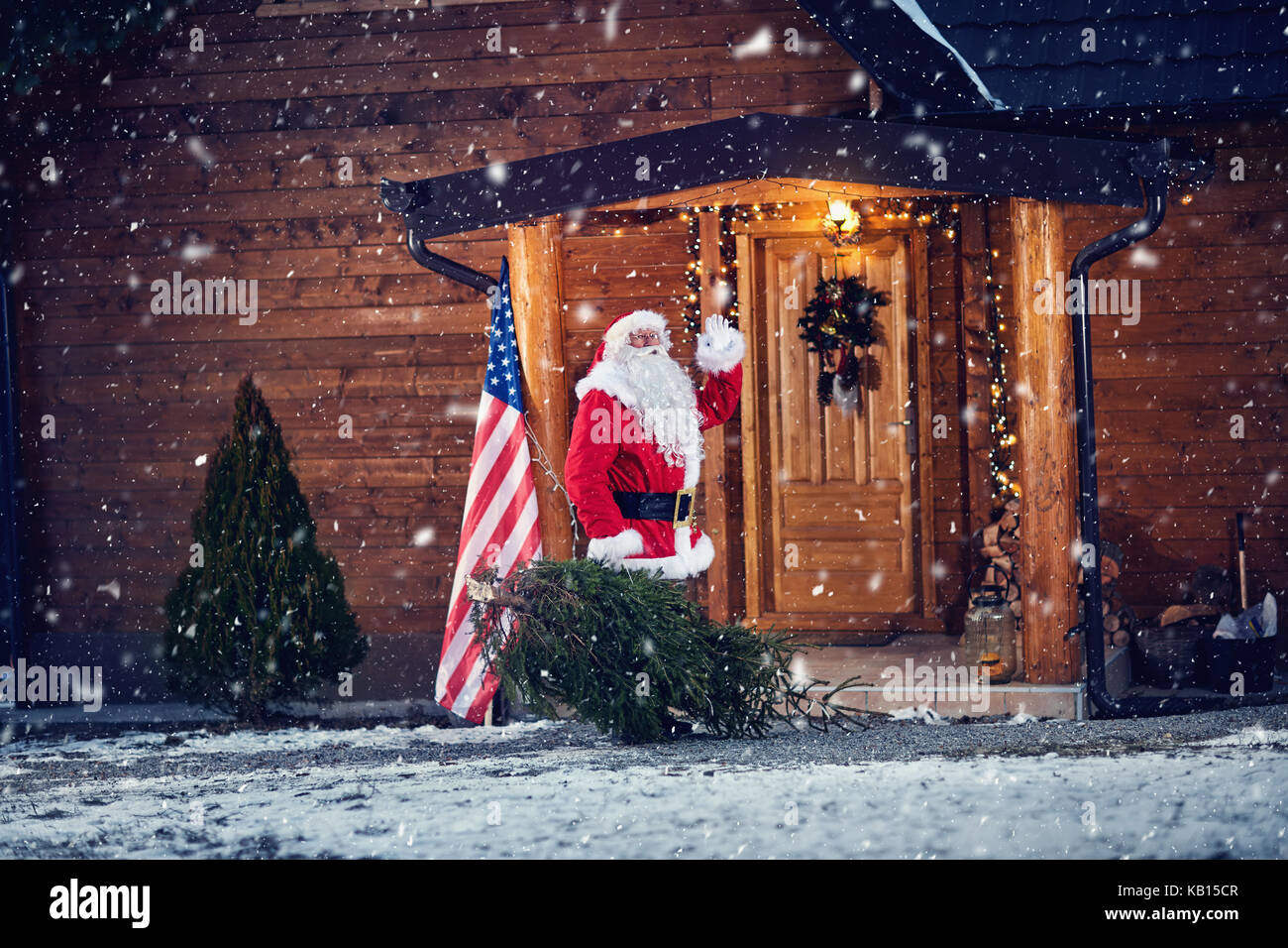 Santa Claus sventolare sul portico anteriore della sua casa in legno e tenuta albero di natale Foto Stock