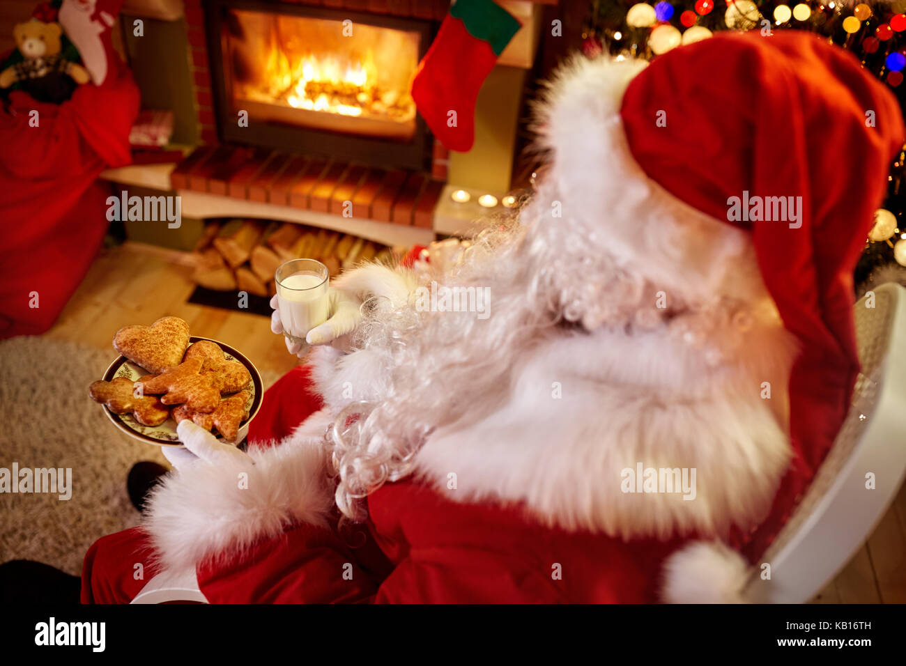 Babbo Natale in un momento di relax a casa in seduta confortevole sedia a dondolo vicino albero di natale il latte da bere e mangiare i biscotti freschi Foto Stock