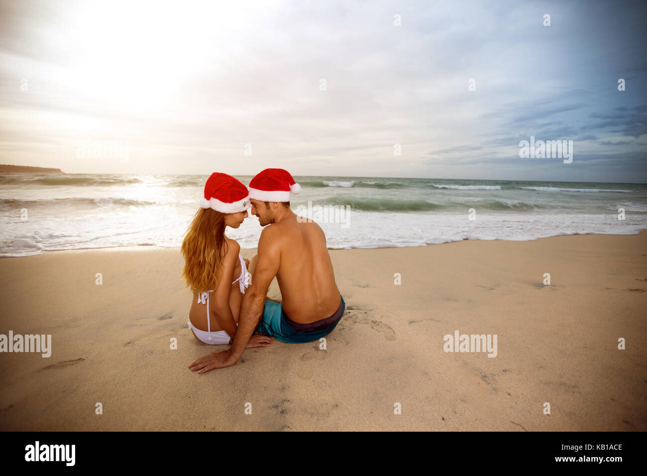 Vista posteriore del giovane con cappelli di Babbo Natale seduto sulla spiaggia sabbiosa Foto Stock