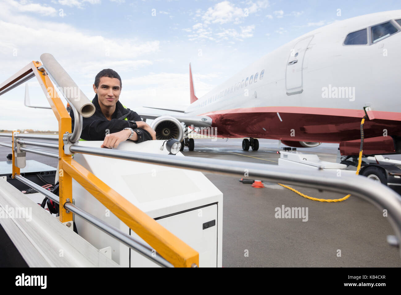 Ritratto di fiducia lavoratore maschile seduto sul trasportatore di bagagli carrello contro aereo all'aeroporto Foto Stock