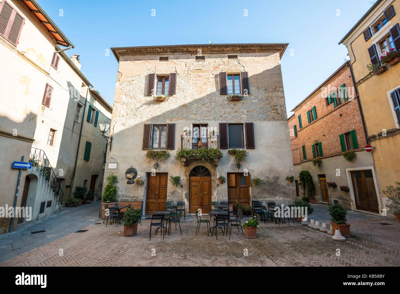 Una piccola piazza in pietra di Pienza con tavoli da caffè, sedie e una bella casa con balcone decorato con fiori Foto Stock