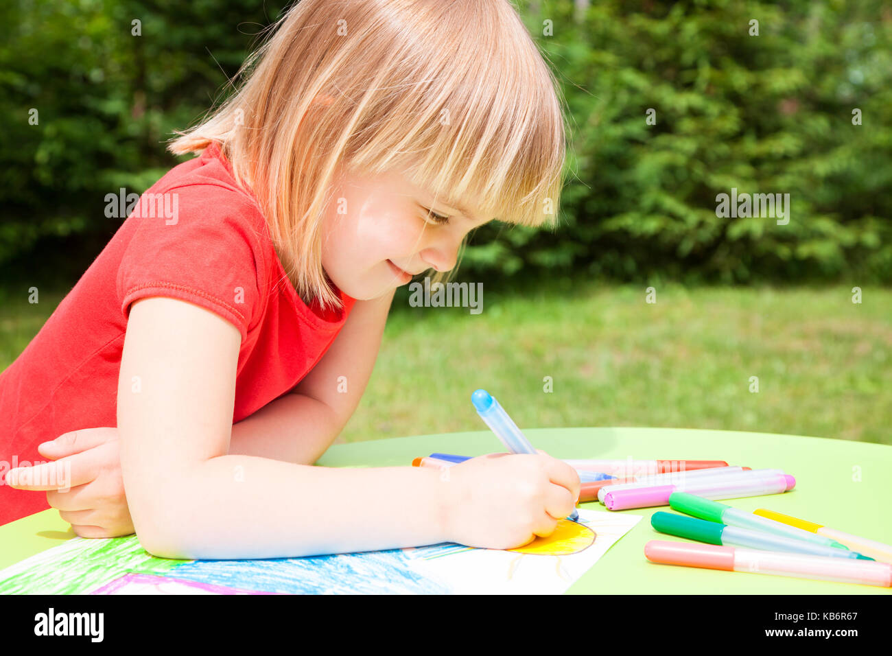 Ritratto di carina ragazza bionda che indossa red tshirt seduti ad un tavolo in un giardino estivo di disegno con pennarelli Foto Stock