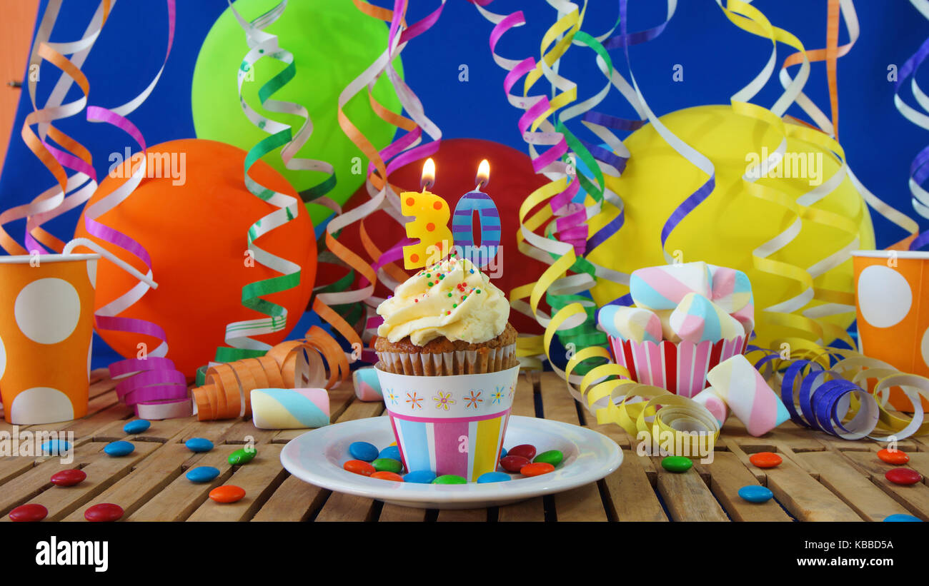 30 compleanno tortina con candele accese sulla tavola in legno rustico con sfondo di palloncini colorati con bicchieri di plastica caramelle sulla parete blu in background Foto Stock