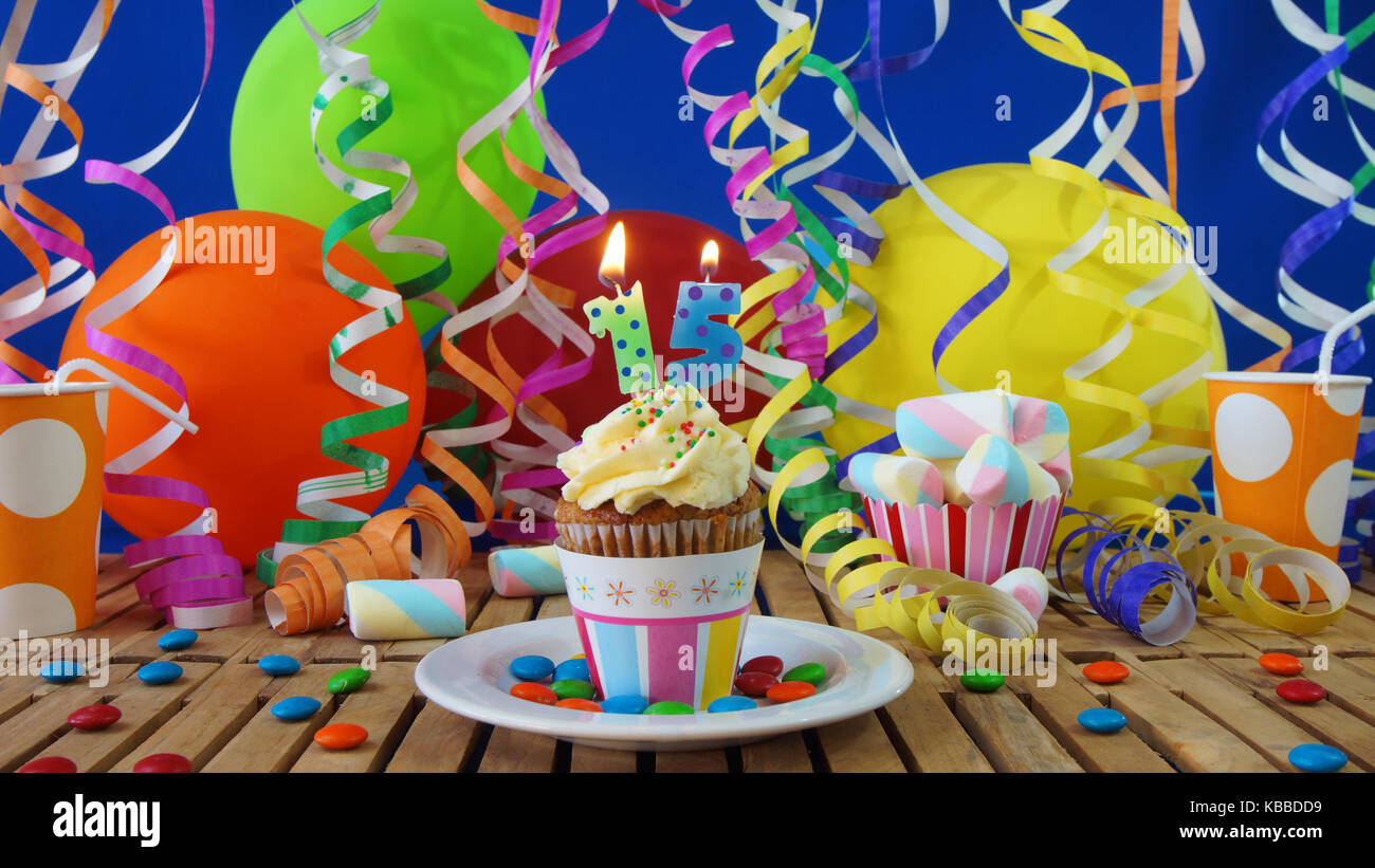 15 compleanno tortina con candele accese sulla tavola in legno rustico con sfondo di palloncini colorati con bicchieri di plastica caramelle sulla parete blu in background Foto Stock