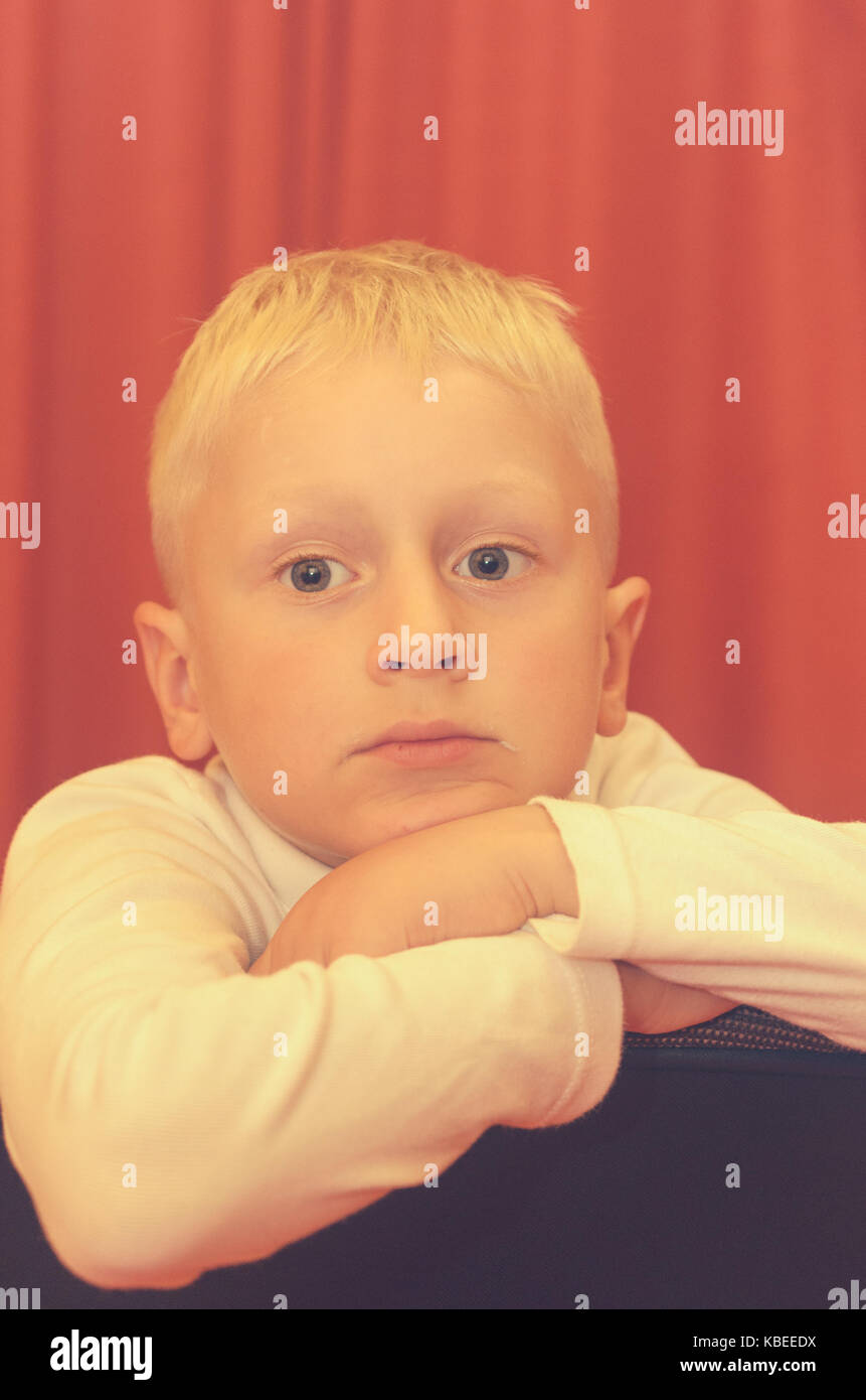 Un ritratto di spalla di un piccolo ragazzo seduto su una sedia con una faccia pensosa Foto Stock