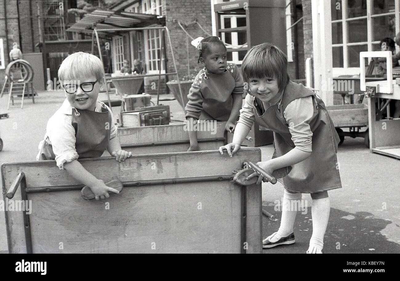 Anni Settanta, foto storiche di tre giovani felici i bambini che indossano tute con le spazzole per pulire un tavolo esterno a un nido su un alloggiamento del consiglio estate in Deptford, a sud di Londra, Inghilterra. Foto Stock