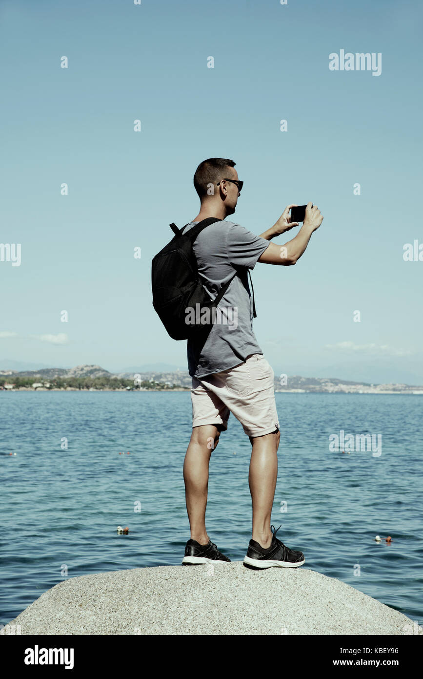 Primo piano di un giovane uomo caucasico portando uno zaino di scattare una foto del mare della costa smeralda, in Sardegna, Italia, con il suo smartphone Foto Stock