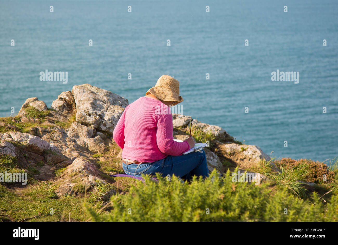 Vista posteriore dell'artista femminile senior in cappello di paglia floppy seduto isolato sulla costa del Regno Unito percorso, schizzo / pittura vista mare. Godere del concetto di pensione. Foto Stock