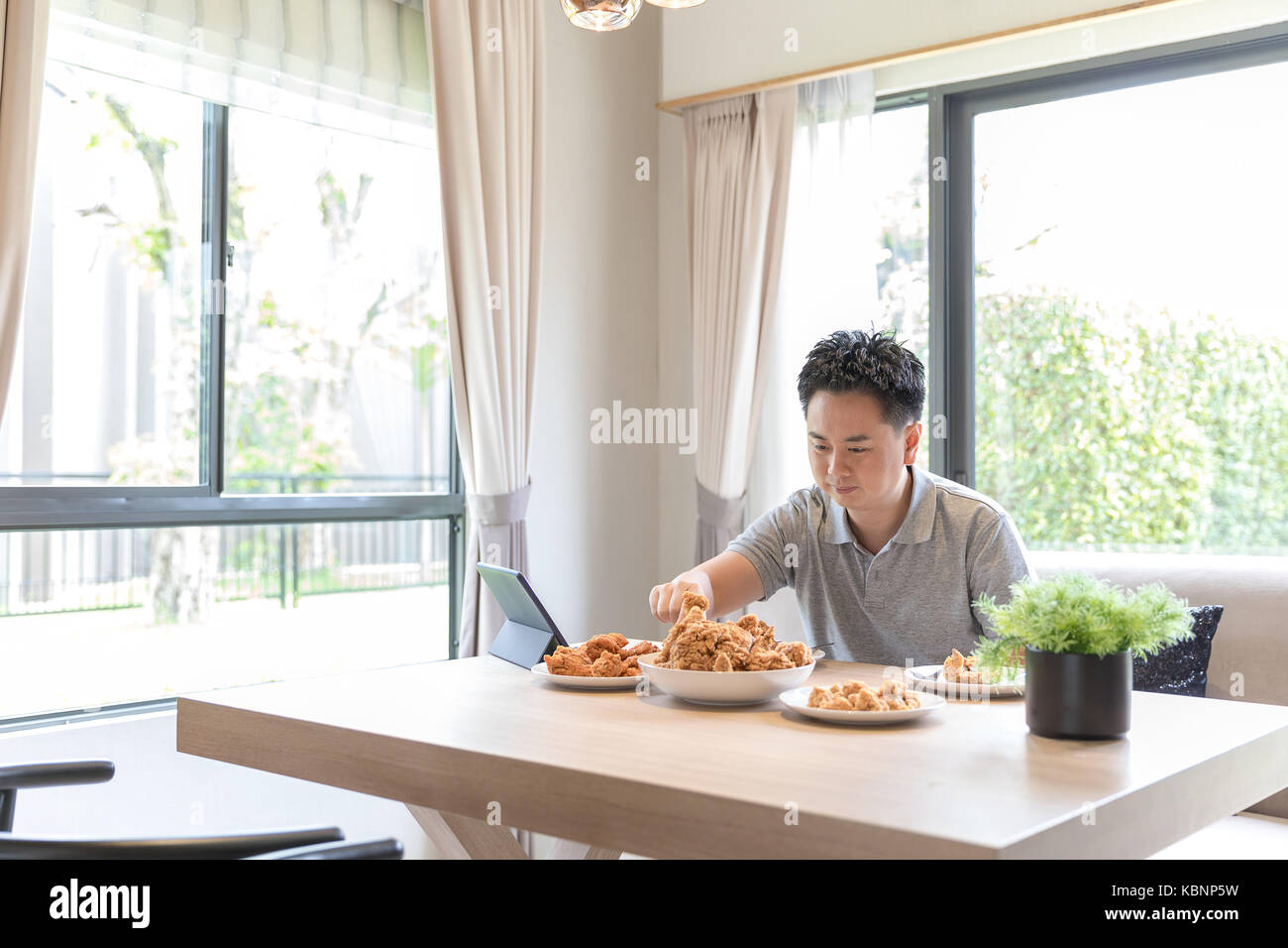 Giovane uomo asiatico di mangiare pollo fritto nel salotto di casa contemporanea per lo stile di vita moderno concetto Foto Stock