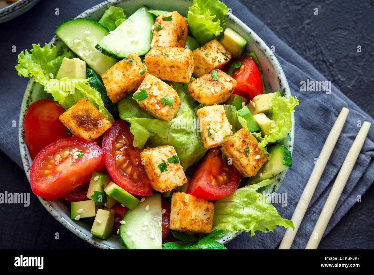 Tofu fritto con insalata di cetrioli e pomodori, avocado e semi di sesamo. in casa asiatica di verdure e insalata di tofu in vaso in ceramica nera su sfondo di pietra Foto Stock