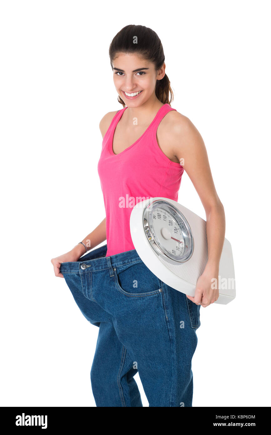 Sorridente giovane donna con peso di scala che mostra il suo vecchio jeans dopo la dieta su sfondo bianco Foto Stock