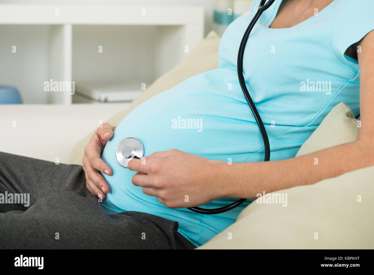 Sezione mediana della donna incinta tenendo uno stetoscopio su stomaco mentre è seduto sul divano di casa Foto Stock