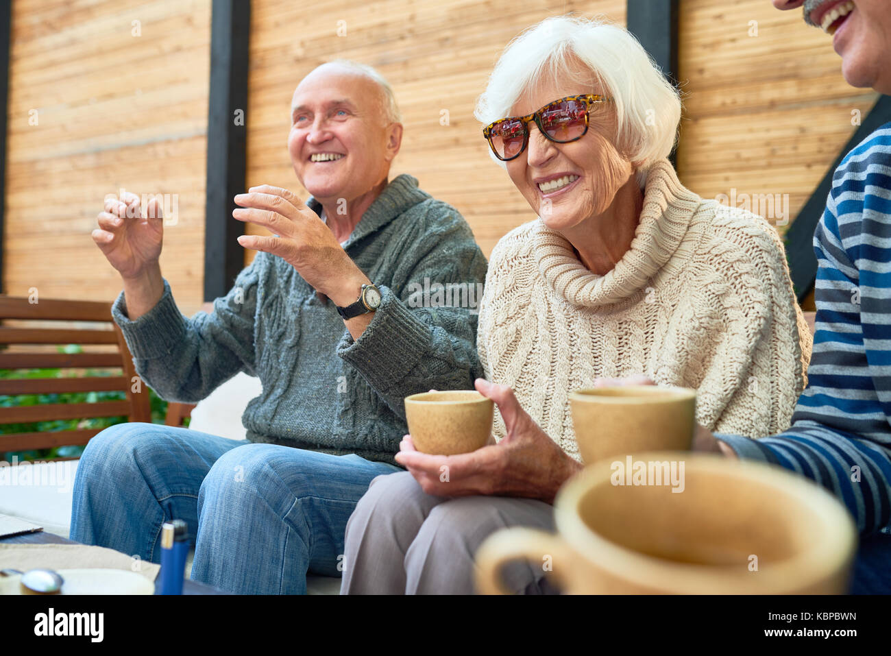 Allegro senior amici radunati insieme al piccolo ed accogliente patio e ricordando storie divertenti dal loro passato, essi indossano Maglioni lavorati a maglia e riscaldamento Foto Stock