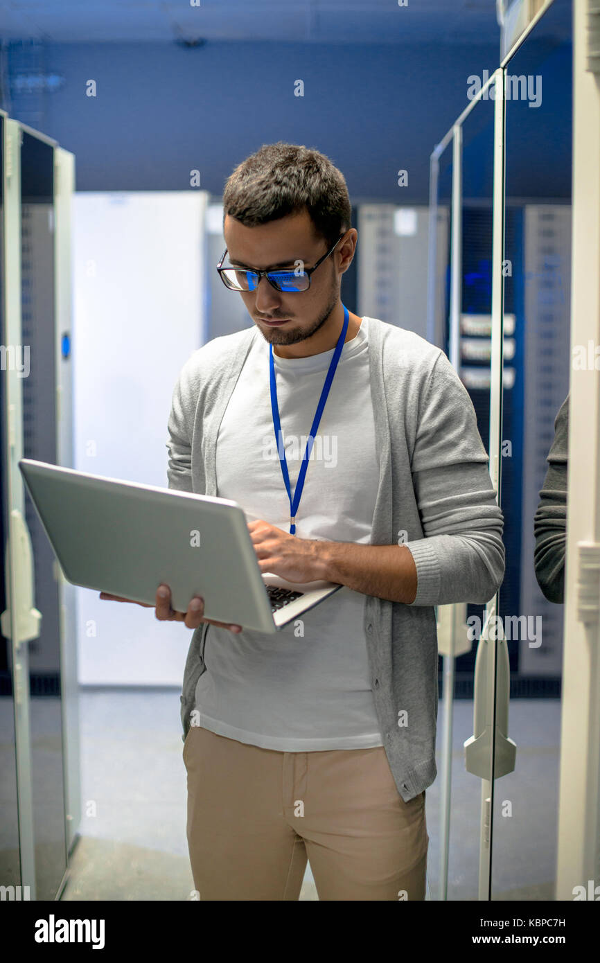 Ritratto di giovane uomo con laptop in piedi da armadi server in dati centro di ricerca Foto Stock