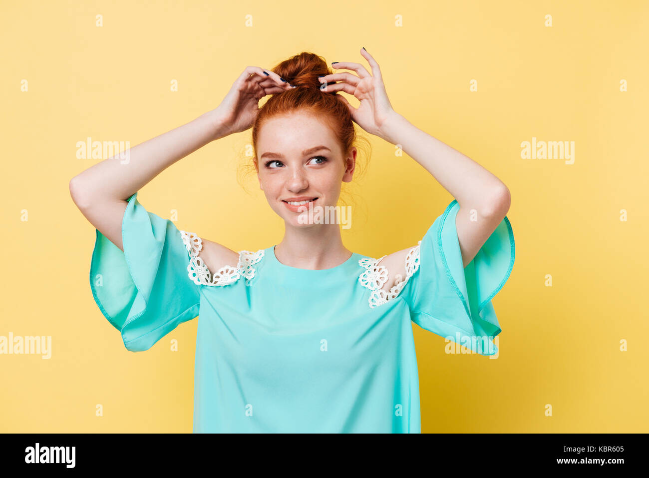 Lo zenzero sorridente donna nel vestire corregge il suo hairstail e guardando lontano su sfondo giallo Foto Stock