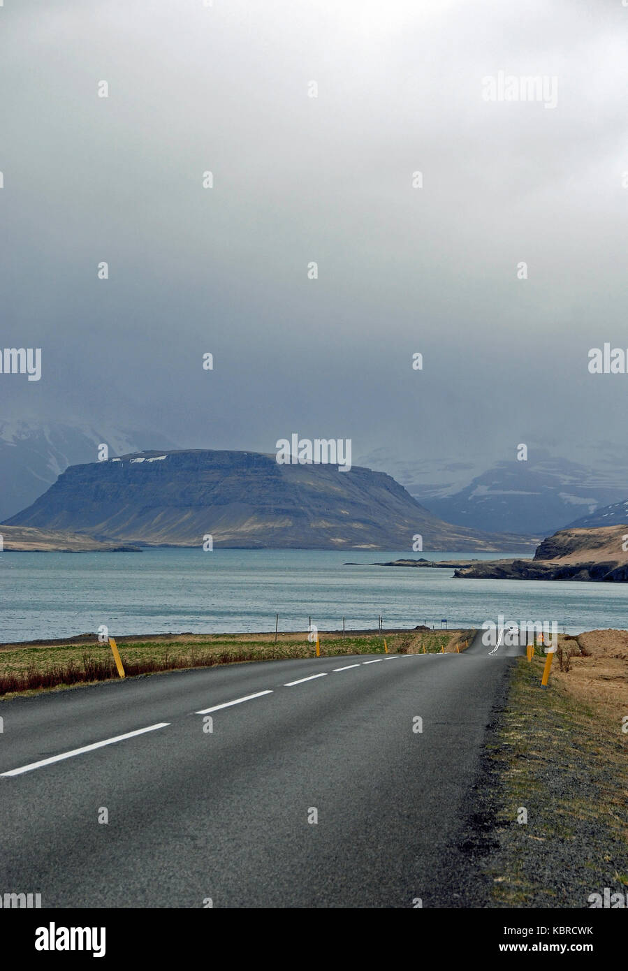 Una vista lungo una strada deserta intorno Hvalfjordur, islandese con montagne in lontananza. Visto sotto un cielo scuro su un molto nuvoloso giorno. Foto Stock