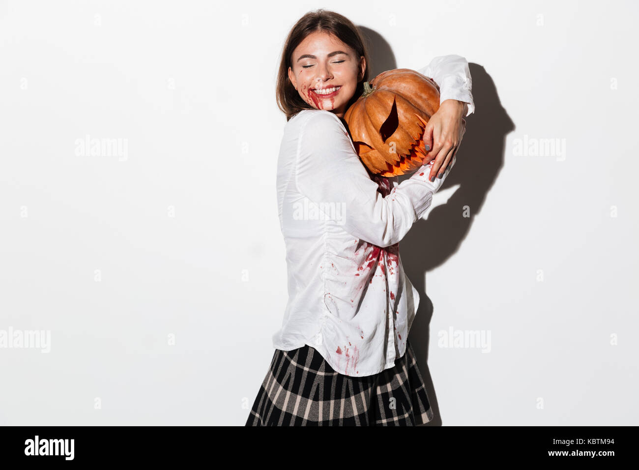 Sorridente carino donna zombie ricoperto di macchie di sangue abbracciando una zucca di Halloween isolate su sfondo bianco Foto Stock