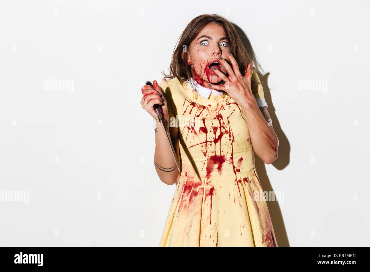 Spaventata terrorizzato zombie donna coperto di sangue in possesso di un coltello e guardando la telecamera isolate su sfondo bianco Foto Stock