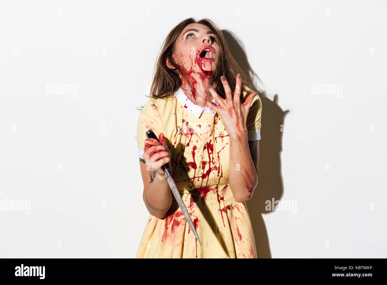 Spaventata mad zombie donna coperto di sangue in possesso di un coltello e guardando in alto isolato su sfondo bianco Foto Stock