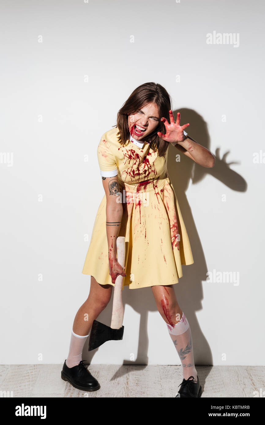 Ful la lunghezza di una donna pazza in zombie make up coperto con macchie di sangue e permanente tenendo un'ascia isolate su sfondo bianco Foto Stock