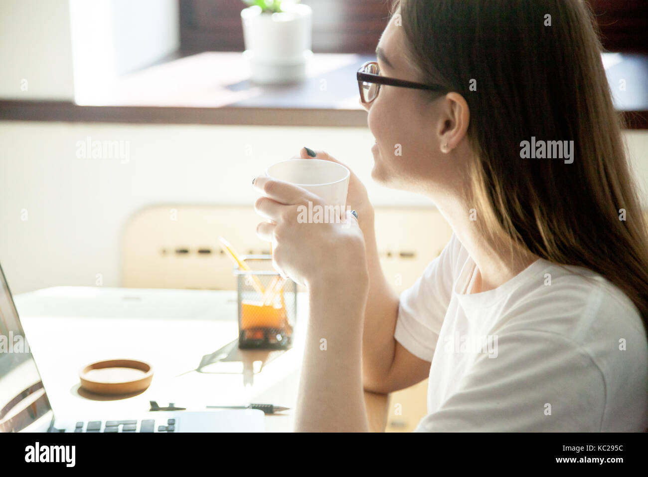 Il dipendente di sesso femminile di prendere una pausa dal lavoro sul  computer portatile, gustando una tazza di tè o caffè alla scrivania sul  posto di lavoro nella piccola casa ufficio. vista