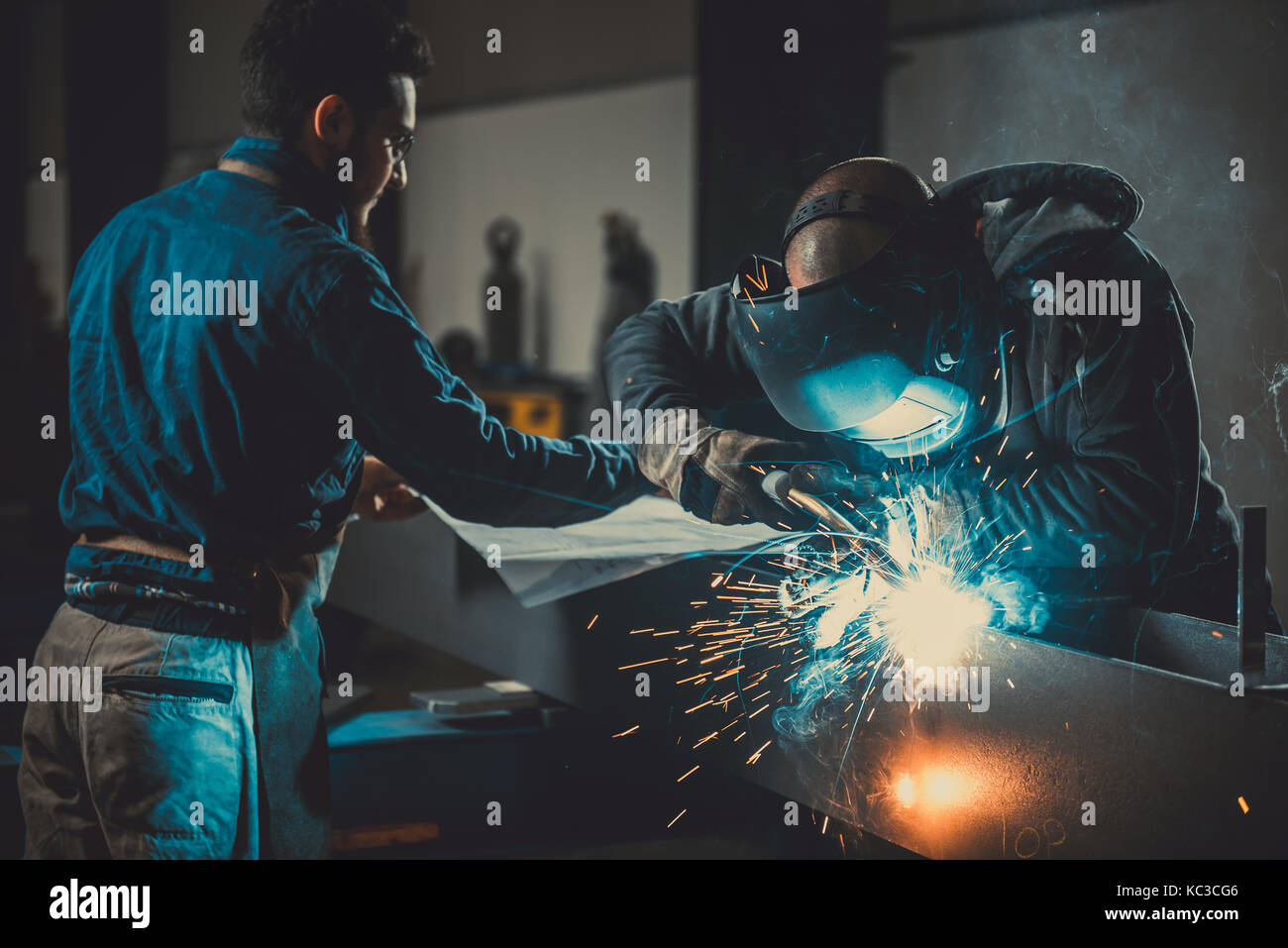 Saldatrice industriale con torcia e casco protettivo nella grande hall di saldatura di profili metallici e in background tecnico blueprint di controllo Foto Stock