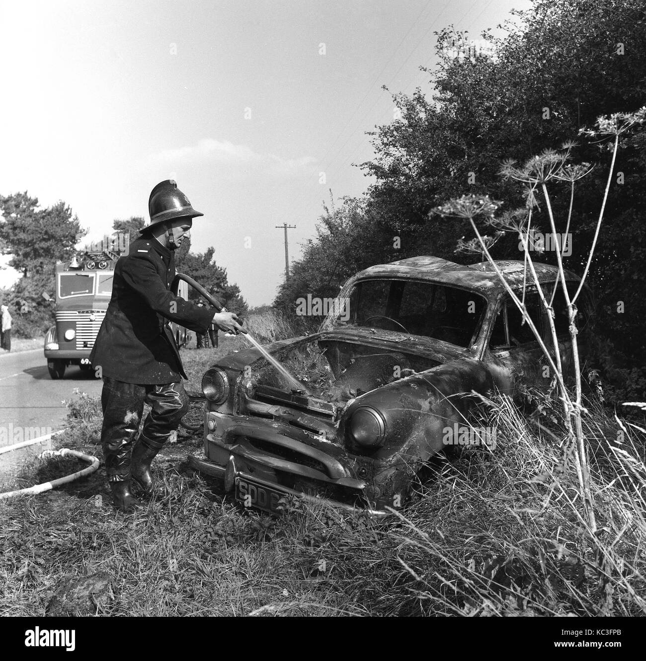 Anni sessanta, storico allarmi antincendio uniforme che indossano il casco e la spruzzatura di acqua in un bruciate le auto di piccole dimensioni su un orlo a lato di una strada di campagna, Inghilterra, Regno Unito. Foto Stock