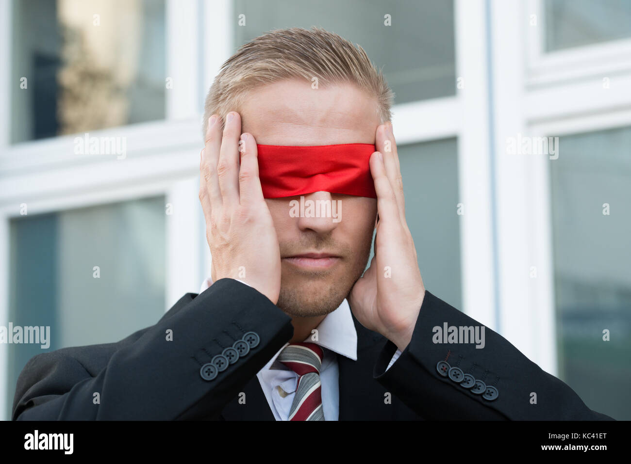 Giovane imprenditore rosso di toccare gli occhi bendati mentre in piedi contro la finestra all'aperto Foto Stock